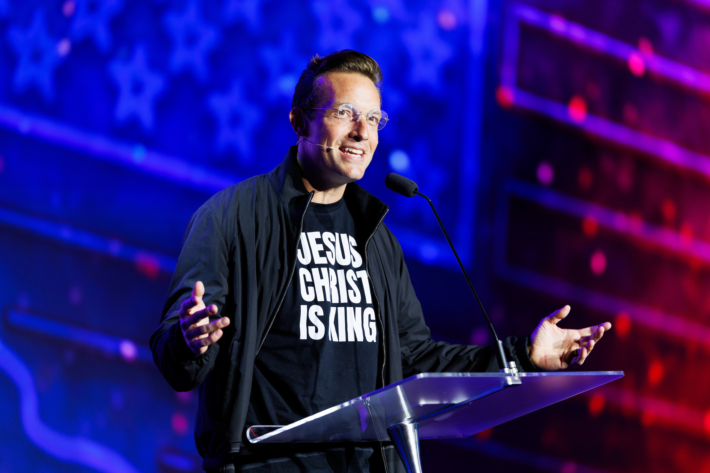 A person wearing a black jacket over a t-shirt that reads "JESUS CHRIST IS KING" stands at a clear podium, speaking into a microphone with hands gesturing outward, against a backdrop of blue and purple lighting.