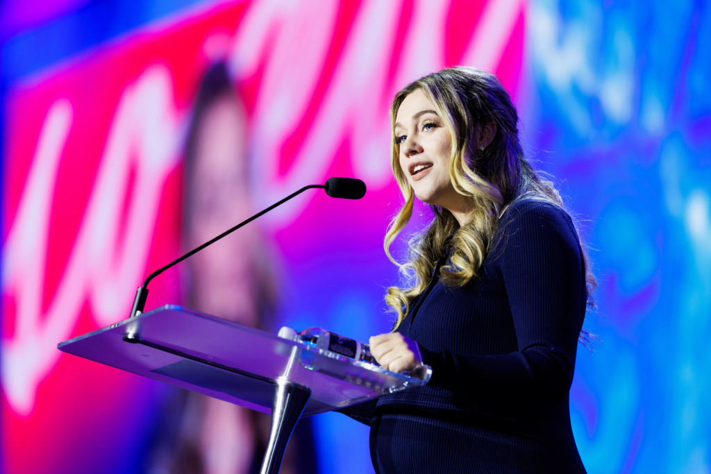 A person with long, wavy hair stands at a transparent podium, speaking into a microphone against a vibrant pink and blue background.