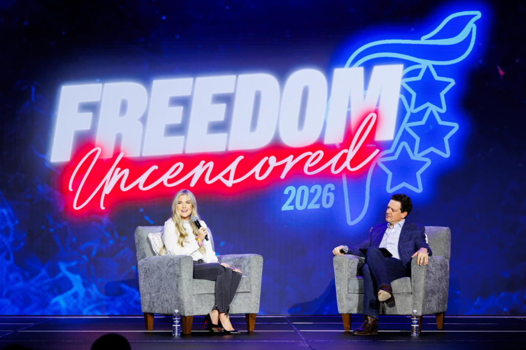 Two individuals sit on stage in armchairs during a panel discussion under a large illuminated sign reading "FREEDOM Uncensored 2026."