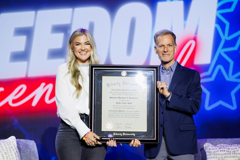 Two individuals stand together holding a framed diploma from Liberty University, with a large "FREEDOM" sign visible in the background.
