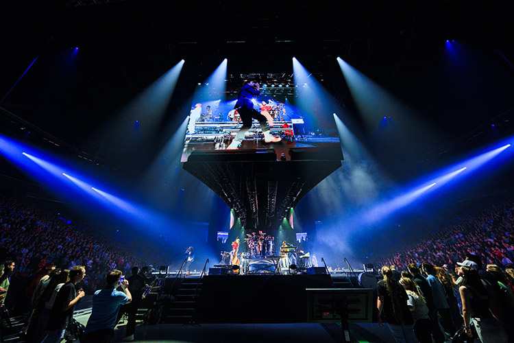A performer leaps on a suspended stage above a live band, illuminated by dramatic blue spotlights in a packed arena.