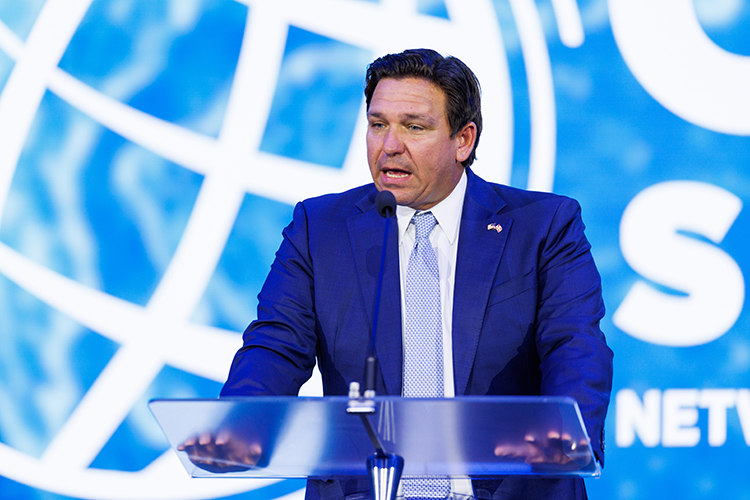A person in a dark suit and tie stands at a podium speaking into a microphone, with a blue background featuring a globe and partial text.