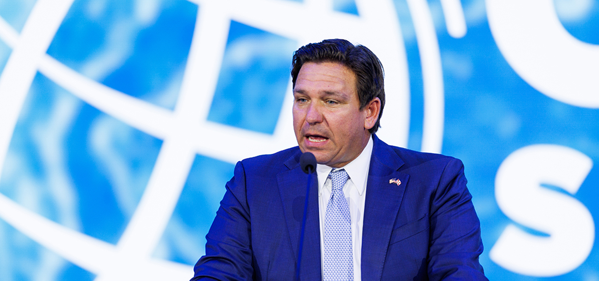 A person in a dark suit and tie stands at a podium speaking into a microphone, with a blue background featuring a globe and partial text.
