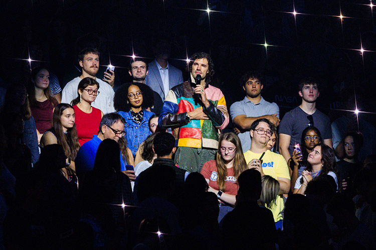 A person in a colorful outfit speaks into a microphone while standing among a group of people in a dark venue illuminated by sparkling lights.