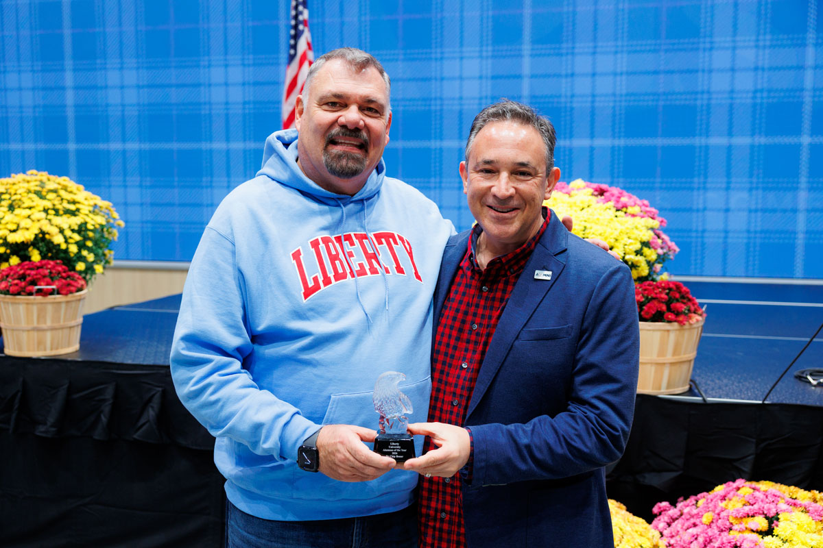 Two individuals stand together on a stage, one holding an award, with colorful flower arrangements and an American flag in the background.