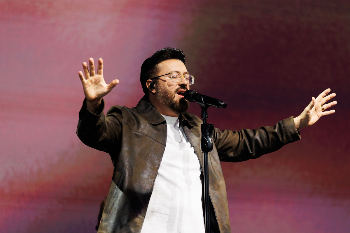 A person with glasses and a beard, wearing a brown jacket over a white shirt, sings into a microphone with arms outstretched against a pinkish-purple backdrop.