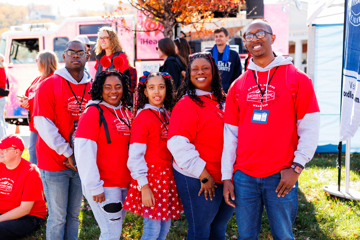 A group of people in matching red hoodies pose together outdoors on a sunny day, with autumn trees and event booths in the background.