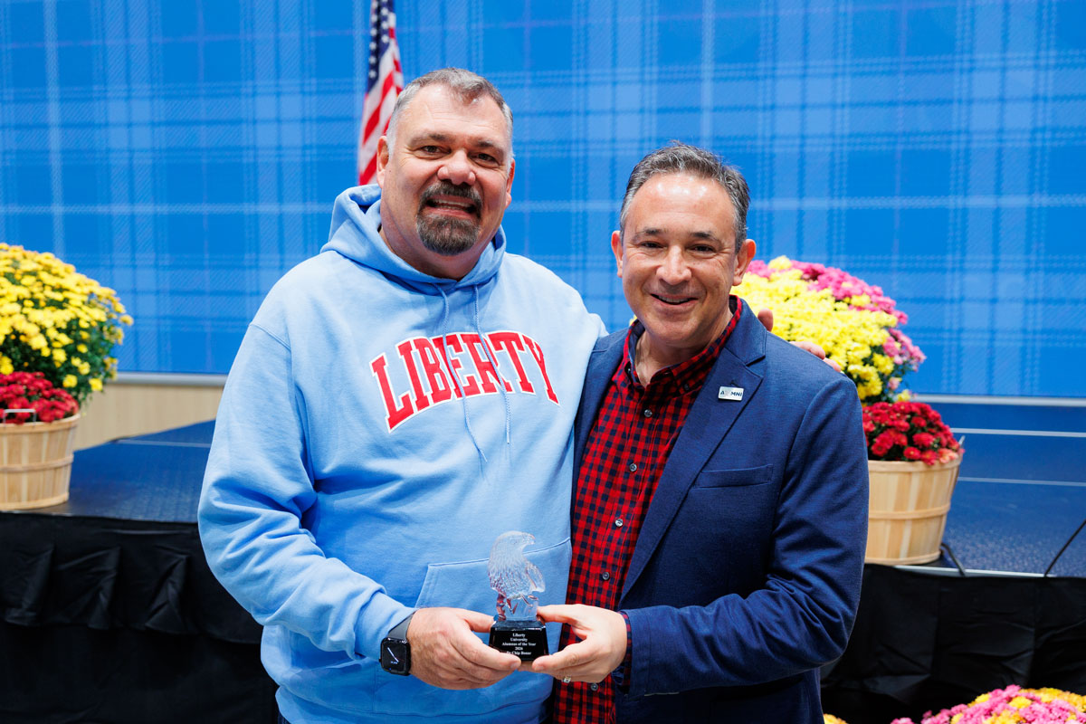 Two individuals stand together on a stage, one holding an award, with colorful flower arrangements and an American flag in the background.