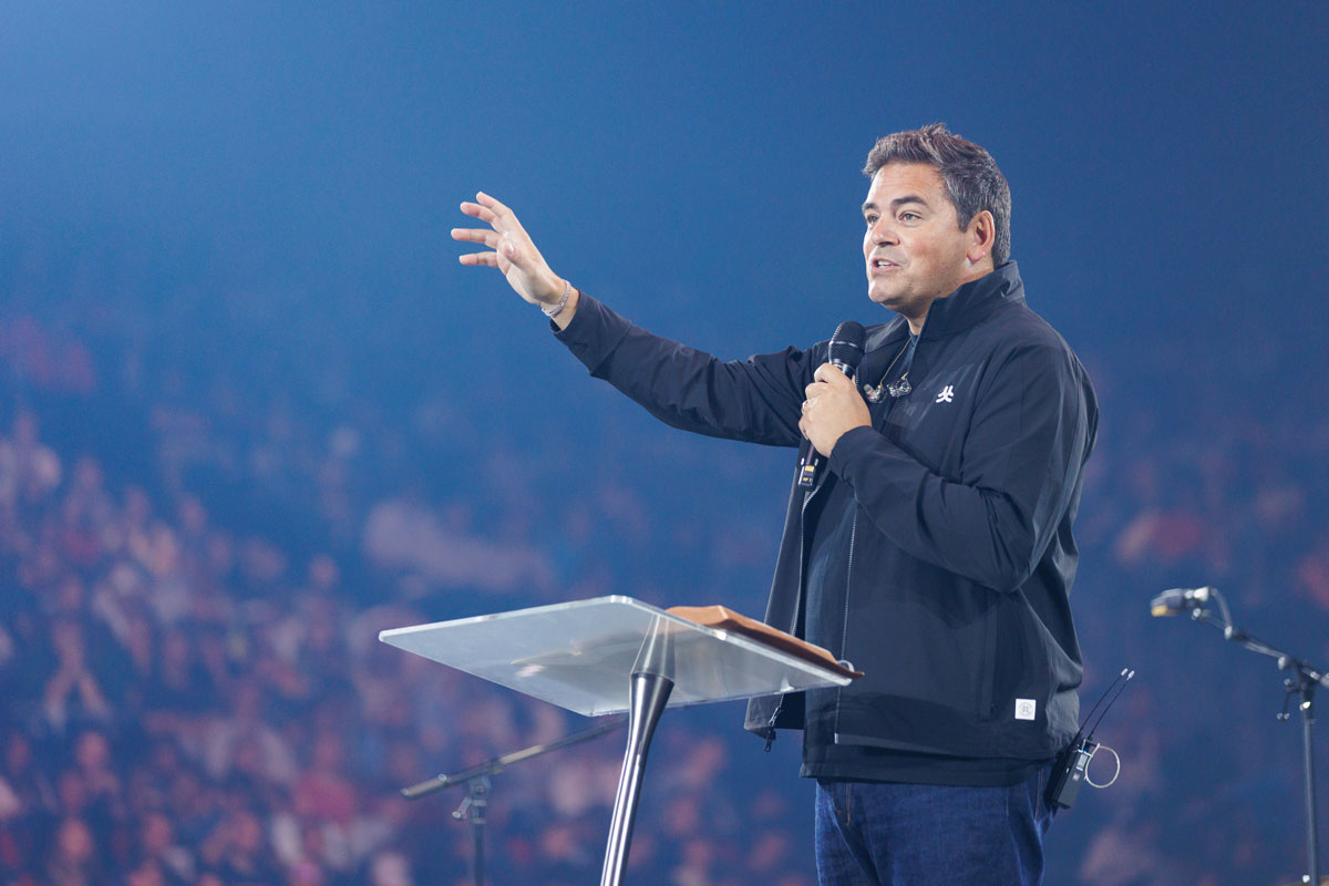 A person stands at a podium, holding a microphone and gesturing outward while speaking to a large audience in a dimly lit venue.