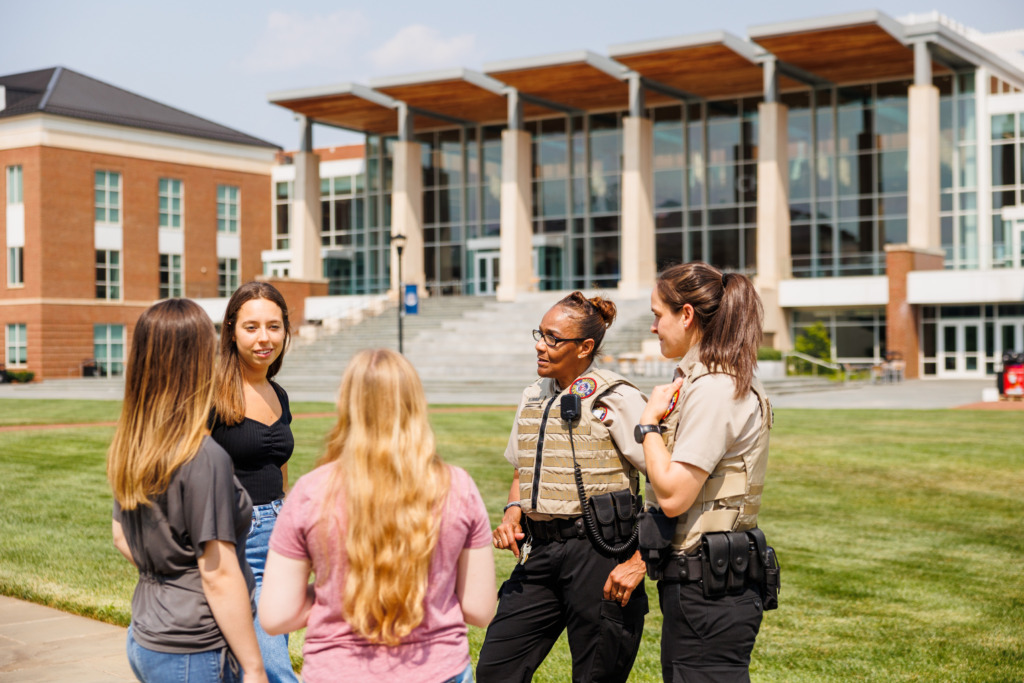 Several individuals, including uniformed officers, are engaged in conversation on a campus lawn in front of modern buildings.