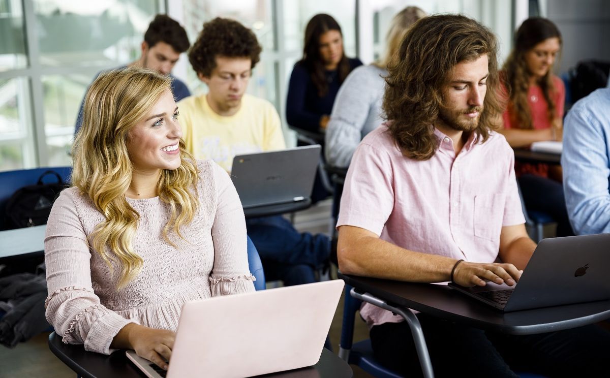 Classroom full of students with laptops, predominantly featuring a blond, smiling, female student.