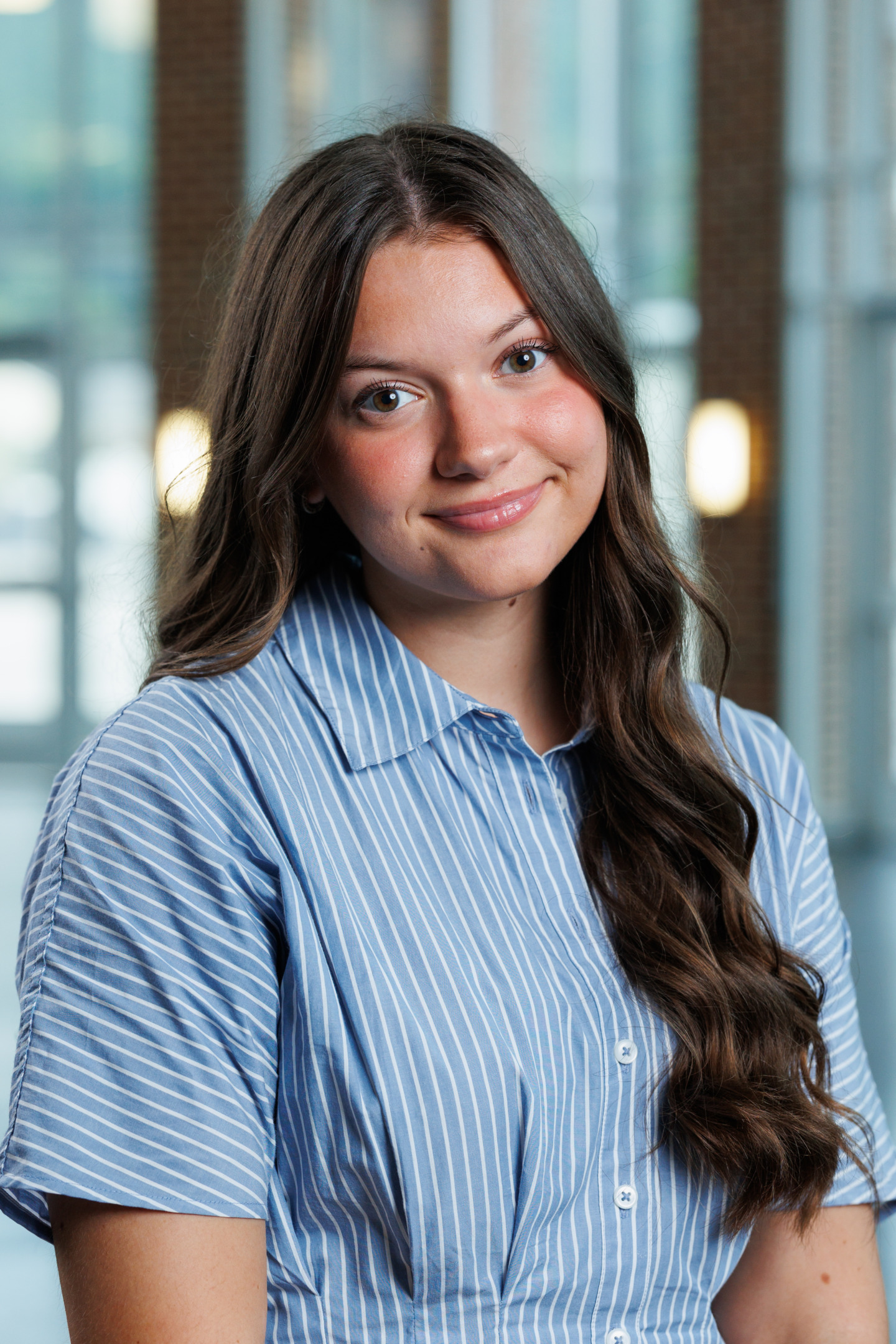 A young person with long brown hair smiles gently while wearing a blue and white striped short-sleeved shirt, set against a softly blurred indoor background.