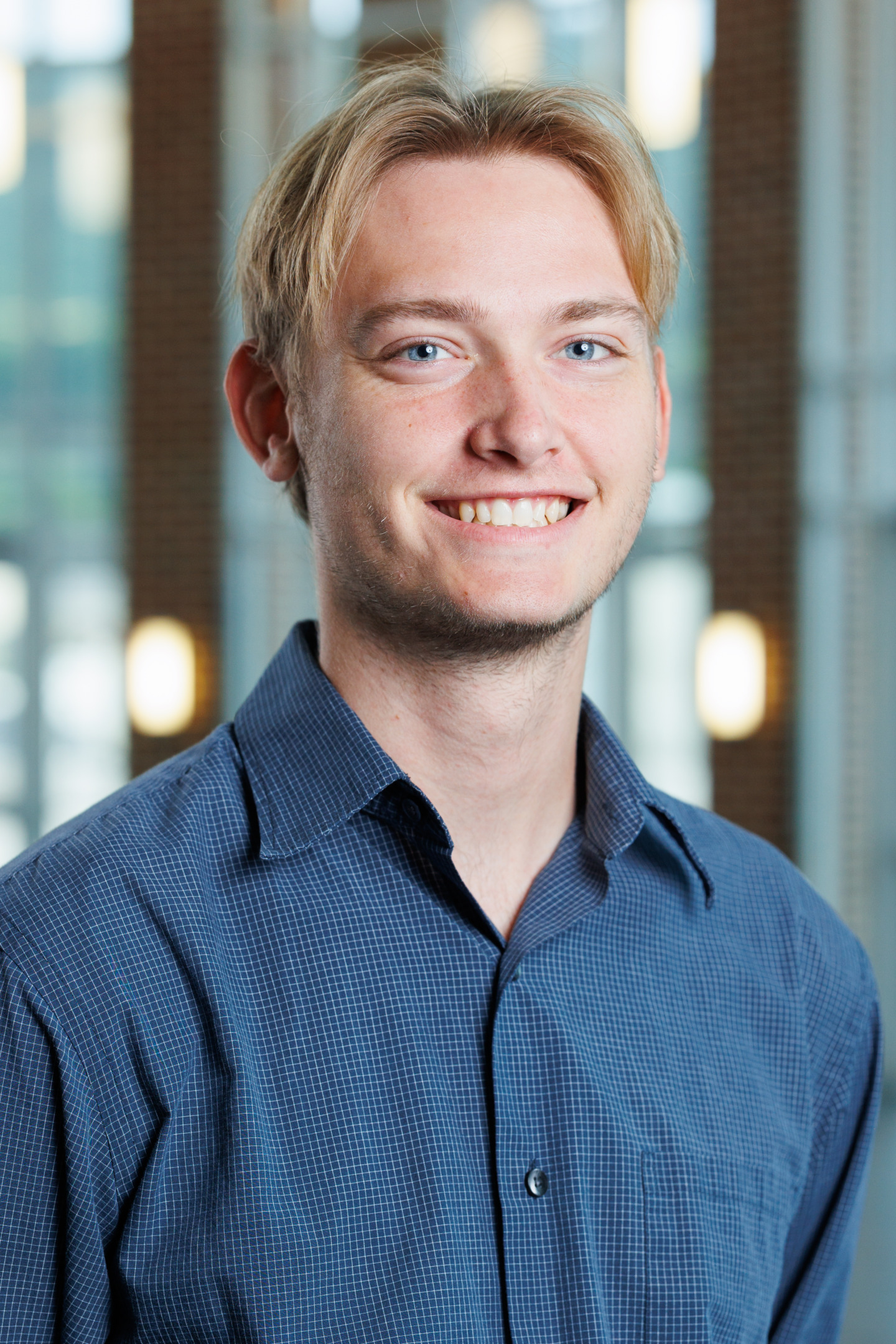 A young person with blond hair and blue eyes smiles warmly while wearing a blue checkered shirt, set against a softly blurred indoor background.