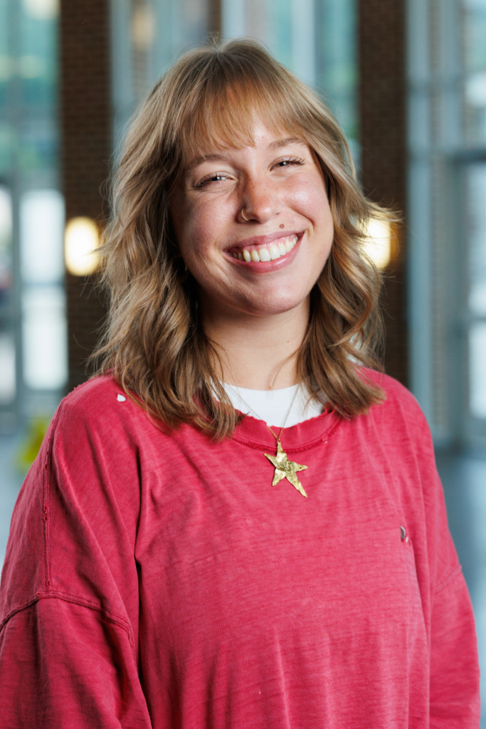 A smiling person with wavy blonde hair and a nose ring wears a red sweatshirt and a gold star necklace, standing indoors with blurred background windows.