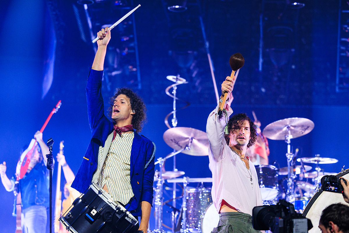 Two percussionists raise their instruments high on stage under blue lighting, with a drum kit and other musicians visible in the background.