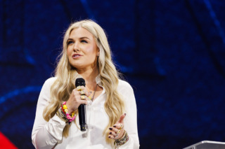 A person with long blonde hair stands on stage holding a microphone, wearing a white shirt and denim pants, gesturing while speaking.